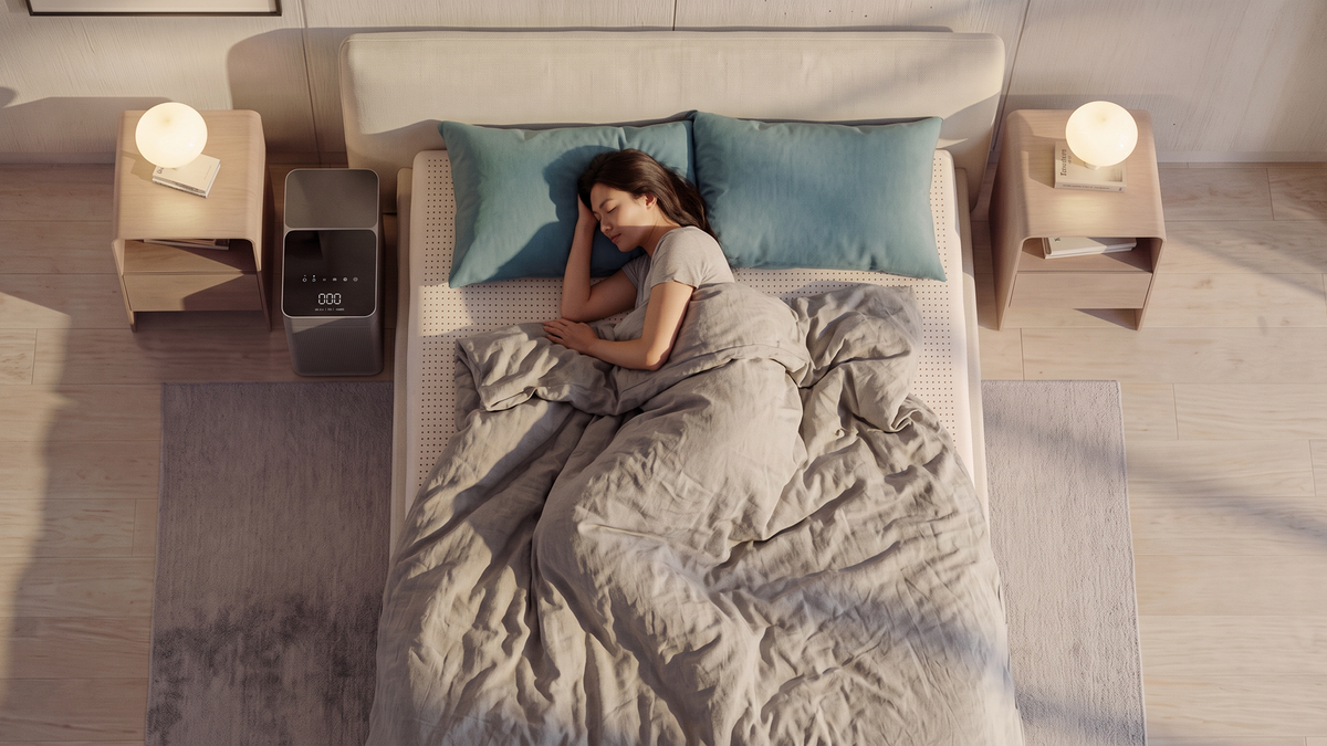 Overhead view of a woman sleeping on a Slowave Dream Pod mattress topper in a bright, sunlit bedroom, with the control unit beside the bed